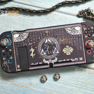 以下が含まれることがあります： 金と白の複雑なパターンと翼を広げた鳥のシンボルが描かれたカスタムスキンを施した黒いニンテンドースイッチ本体。本体には金色のZが刻まれたカスタムボタンが2つ付いている。