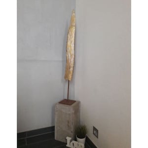 Op de afbeelding: Een hoog, dun, lichtbruin houten beeld met een gouden afwerking staat op een betonnen sokkel. Het beeld is gemonteerd op een metalen staaf. De sokkel bevindt zich in een hoek van een kamer met een witte muur en een zwarte tegelvloer.