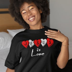 May include: Black crew neck t-shirt with the words "Jesus is Love" printed on the front. The design features four heart shapes, alternating between red and white. The person in the image is smiling.