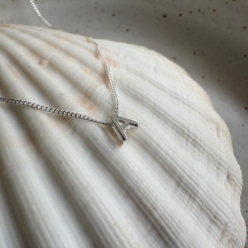 May include: A silver necklace with a small letter A pendant. The necklace is on a white seashell background.