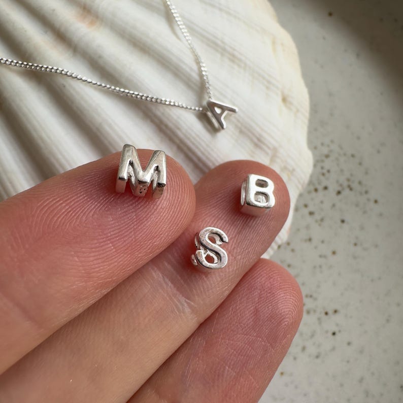 May include: Close-up of silver letter charms, including the letters A, M, B, and S. The charms are on a white surface and a hand is holding them.