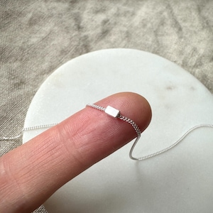 May include: A delicate silver necklace with a small rectangular pendant. The pendant is attached to a fine chain. The jewellery is displayed on a finger, highlighting its minimalist design and subtle elegance.