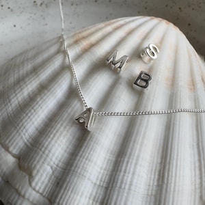 May include: A silver necklace with a silver letter A charm. The necklace is on a white seashell with other silver letter charms, including M, S, and B.