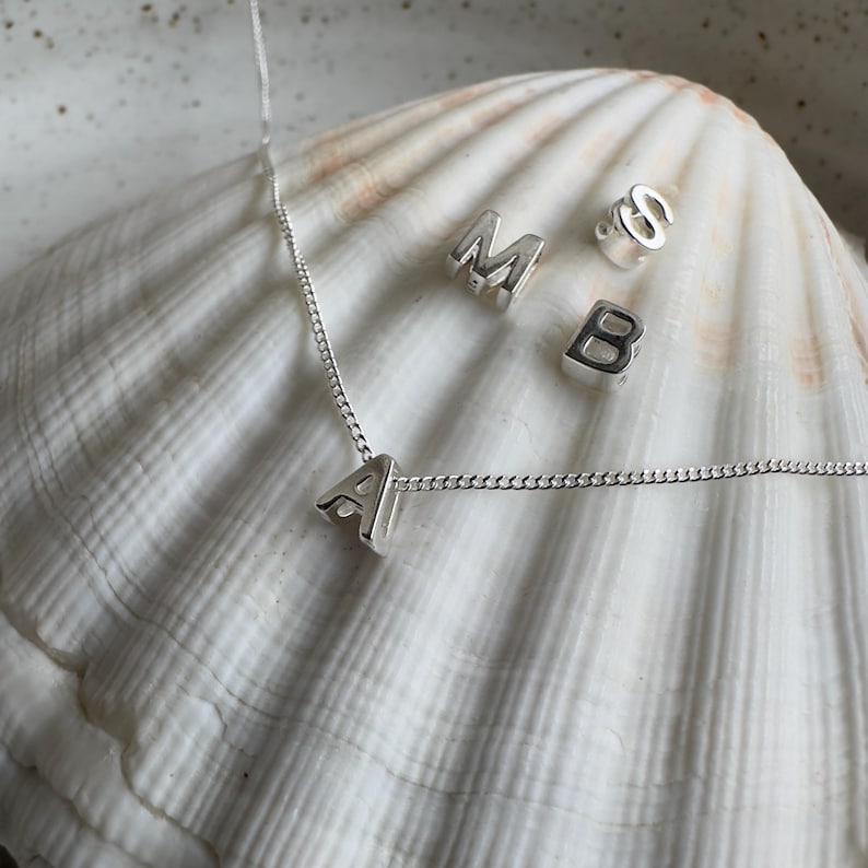 May include: A silver necklace with a silver letter A charm. The necklace is on a white seashell with other silver letter charms, including M, S, and B.