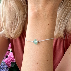 Puede incluir: Un delicado brazalete de cadena plateada con una sola cuenta de vidrio de mar verde claro.