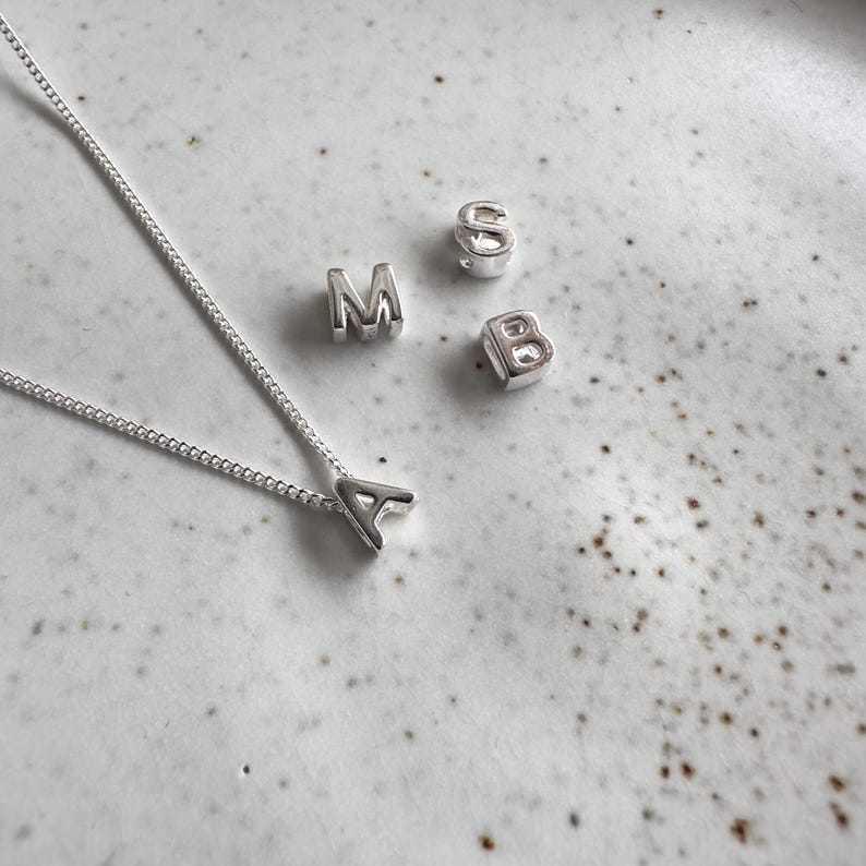 May include: A silver necklace with a silver letter A charm. There are also silver letter charms for the letters M, S, and B.