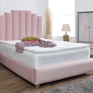 May include: A pink velvet bed frame with a tall headboard featuring vertical panels. The bed has a white mattress and a white and grey patterned topper. The bed is in a bedroom with a white wall and a wooden cabinet.