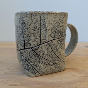 May include: A set of three ceramic mugs with a speckled, matte finish. The mugs are in shades of cream, light gray, and dark gray. Each mug has a simple, minimalist design and a rounded handle.