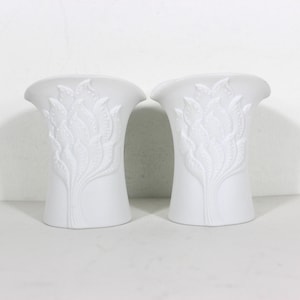 May include: A pair of white ceramic vases with a raised floral design. The vases are tall and slender with a wide mouth.
