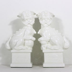 May include: A pair of white ceramic foo dog statues, each with a raised paw and a detailed face. The statues are sitting on square bases.