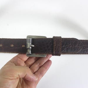 May include: A brown leather belt with a silver buckle. The belt has a textured surface and is stitched with a decorative pattern.