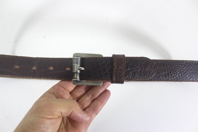 May include: A brown leather belt with a silver buckle. The belt has a textured surface and is stitched with a decorative pattern.