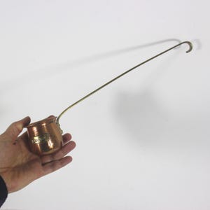 May include: A copper ladle with a long, thin handle. The ladle has a small, round bowl with a label that reads "BRANDY".
