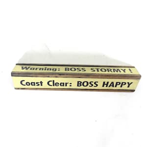 May include: Wooden desk sign with the text "Warning: BOSS STORMY!" and "Coast Clear: BOSS HAPPY" in black lettering. The sign has a light yellow top and bottom with a dark brown wooden center.