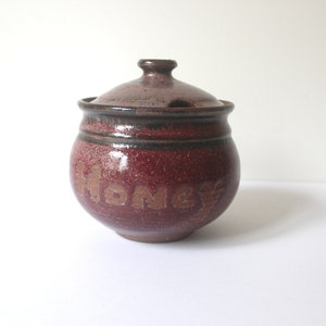 May include: A round ceramic honey pot with a lid. The pot is a deep red colour with brown accents and the word "HONEY" is written on the side in a lighter brown colour. The lid has a small knob.