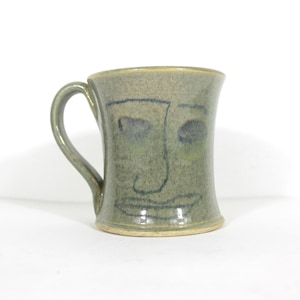 May include: A handmade ceramic mug with a whimsical face design. The mug is a muted green color with a curved handle. The face is drawn with dark blue lines, featuring eyes, a nose, and a smiling mouth. The mug has a slightly flared shape.