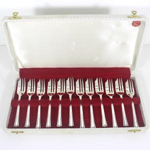 May include: A set of twelve silver forks in a white and red lined case. The forks are arranged neatly in a velvet-lined compartment. The case has a textured exterior and gold-colored clasps.