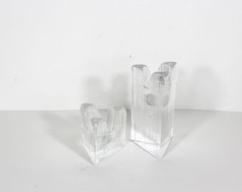May include: Two clear glass candle holders of varying heights, each featuring a triangular base and a textured, vertically ribbed surface. The tops of the candle holders have a notched, multi-pointed design, creating a distinct silhouette for each piece.