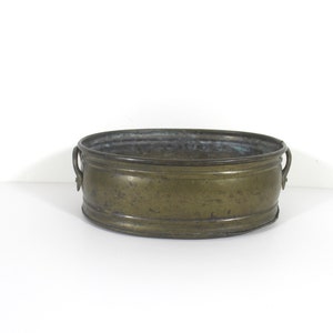 Peut inclure: Un pot en laiton avec deux poignées. Le pot est de forme ovale et a un aspect usé et vieilli.