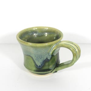 May include: A green ceramic mug with a flared rim and a unique, sculpted handle. The mug has a light green glaze with a blue and white marbled pattern.