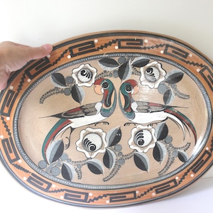 May include: A large oval ceramic platter with a brown, black and white geometric border. The centre of the platter features a painted design of two birds with long tails, surrounded by white flowers and green leaves.