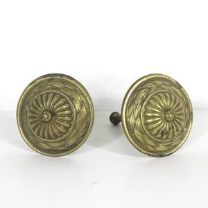 Peut inclure: Deux boutons de porte anciens en laiton. Chaque bouton présente un motif circulaire avec un motif floral central entouré d'une bordure de feuilles détaillée. Les boutons sont de couleur laiton vieilli et chaleureux.
