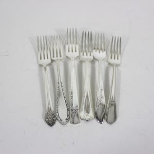May include: Six silver forks with ornate designs on the handles. The forks are arranged in a row, with the handles facing the viewer.