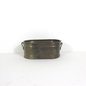 May include: A small oval brass planter with two handles. The planter is made of shiny, aged brass.