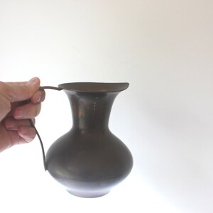 Pode incluir: Um jarro de metal castanho escuro com uma base larga e arredondada e uma borda alargada. O jarro tem uma pega simples e curva.