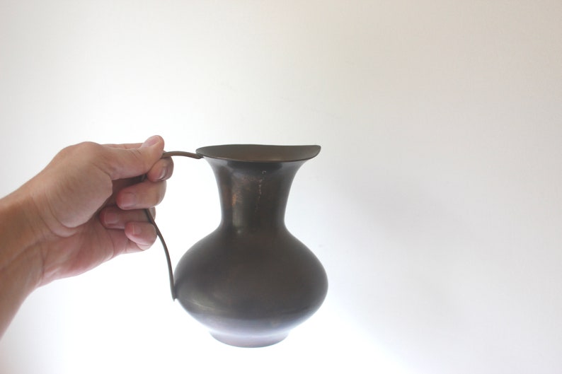Pode incluir: Um jarro de metal castanho escuro com uma base larga e arredondada e uma borda alargada. O jarro tem uma pega simples e curva.