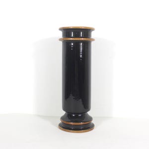 May include: A tall, black ceramic vase with a glossy finish. The vase has a cylindrical body with a flared base and a slightly wider top. It features a brown band at the top and base, adding a contrasting accent. The vase is set against a white background.