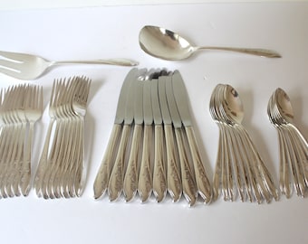May include: A collection of silver-colored flatware, including forks, knives, and spoons, arranged on a white surface. The set includes multiple forks, knives, and spoons, along with a serving fork and spoon. The silverware has a decorative floral pattern.