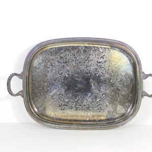 May include: A silver serving tray with ornate embossed designs and two handles. The tray is oval shaped and has a slightly raised rim.