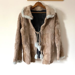 May include: A tan faux fur jacket with a light blue trim on the collar and cuffs. The jacket has a tie closure with light blue pom-poms. The jacket is hanging on a wooden hanger.