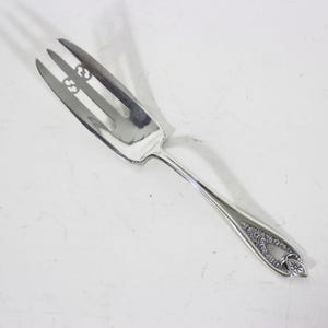 May include: A silver serving fork with three tines and a long handle. The tines have decorative cutouts. The handle features an ornate floral design near the end. The fork is likely made of stainless steel.
