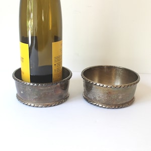 May include: Two silver-toned wine bottle coasters with a rope-like design around the top and bottom edges. One coaster holds a wine bottle. The coasters have a vintage, antique appearance.