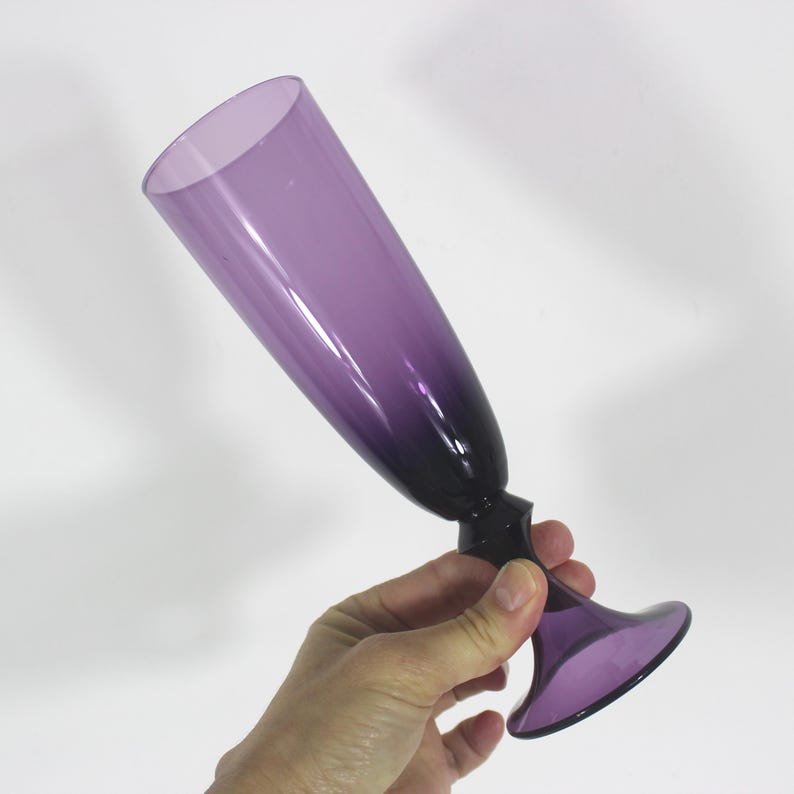 May include: A tall, purple glass champagne flute with a flared base. The glass has a gradient color, fading from a lighter purple at the top to a darker purple at the base.