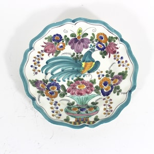 May include: A white ceramic plate with a blue rim and a colorful floral design featuring a peacock in a vase. The flowers are pink, yellow, orange, and purple.