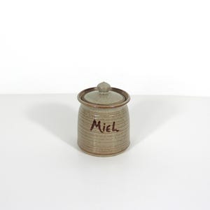 May include: A small, tan ceramic jar with a lid. The jar has a ribbed texture and the word "Miel" is written in brown script on the front. The jar is likely used for storing honey or other food items.