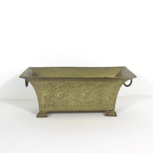 May include: A brass planter with an ornate design and two handles. The planter has four feet and is rectangular in shape.