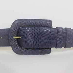 May include: A dark navy blue belt with a textured, lizard-skin-like pattern. The belt features a matching dark navy blue buckle covered in the same textured material, secured by a thin, metallic gold-toned prong and two matching belt loops.