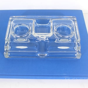 May include: A clear glass inkwell with three compartments. The inkwell is rectangular in shape and made of thick glass.