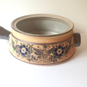 May include: A ceramic casserole dish with a tan exterior and a dark grey handle. The dish features a floral and bird design in blue, green, and grey. The interior is light grey.