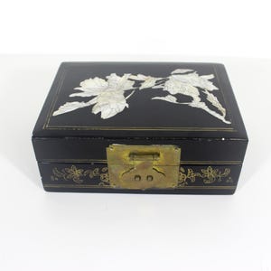 May include: Black rectangular jewelry box with a floral design on the lid. The box has a brass clasp and floral accents along the bottom edge. The floral design on the lid is white.