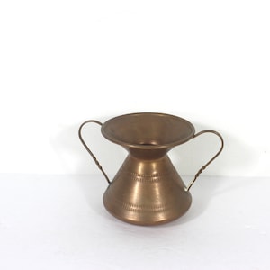 May include: A small, brass vase with two handles. The vase has a wide, flared top and a narrow, rounded bottom. The surface of the vase is textured.