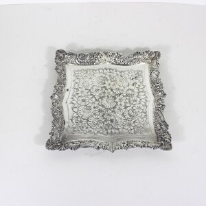 May include: A square silver-toned metal tray featuring an intricate embossed floral pattern across the centre surface. The edges of the tray are decorated with a detailed, raised scrollwork and leaf border design, creating a textured frame.