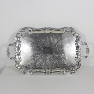 May include: A rectangular silver-toned serving tray featuring ornate, scalloped edges and two decorative handles. The surface of the tray is adorned with intricate, embossed floral and scrollwork patterns that cover the entire central area.