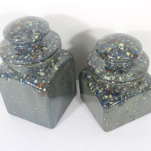 May include: Two ceramic jars with lids. The jars are square-shaped with a speckled blue-grey glaze. The lids are round and stacked, also with the speckled glaze. The jars have a rustic, handmade appearance.