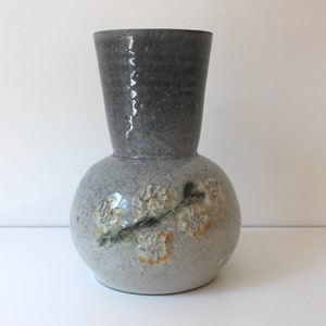 May include: A handmade ceramic vase with a rounded base and a cylindrical neck. The vase is mainly grey with a darker grey neck. Floral relief details in white and yellow decorate the base. A unique decorative item.