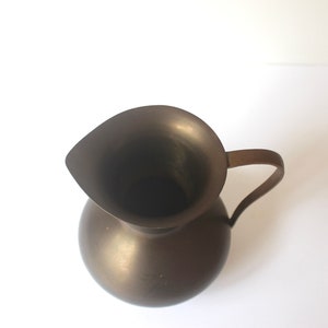 Pode incluir: Um jarro de metal castanho escuro com uma base larga e redonda e uma parte superior em forma de sino. O jarro tem uma &uacute;nica pega.